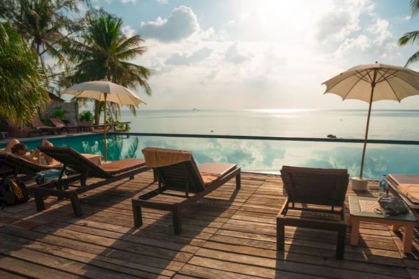 A serene poolside scene with lounge chairs, umbrellas, and a stunning view of the ocean and sky, under the shade of palm trees, on a sunny day.