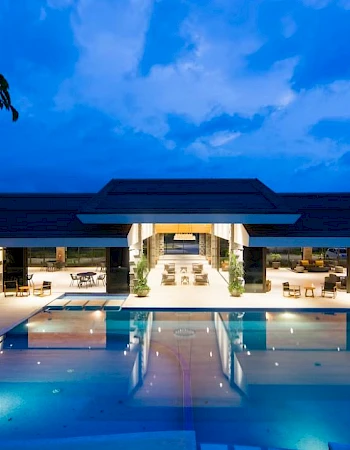 An illuminated outdoor swimming pool area in front of a large, modern building with open architecture under a twilight sky, featuring lounge areas.
