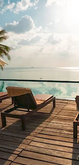 A serene poolside setting with lounge chairs, umbrellas, palm trees, and a stunning view of the ocean under a bright sky.