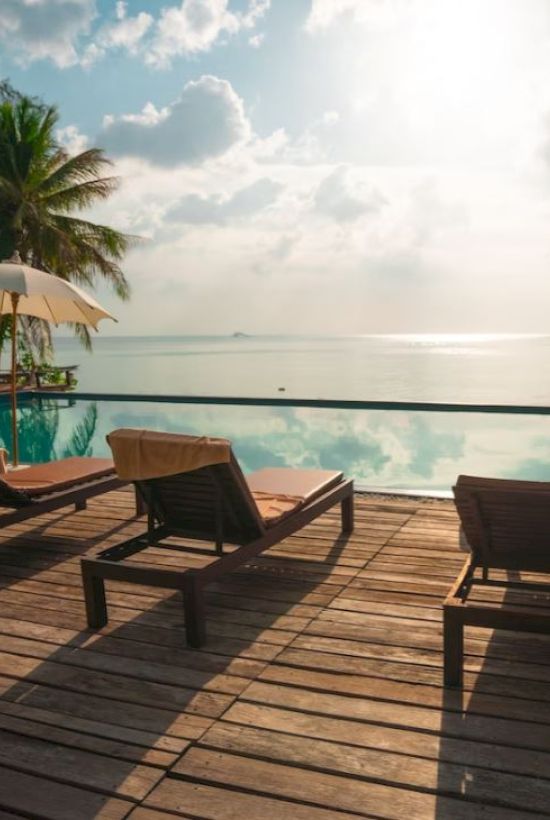 Lounge chairs and umbrellas by a serene pool, overlooking the ocean with palm trees and a sunlit sky in the background.