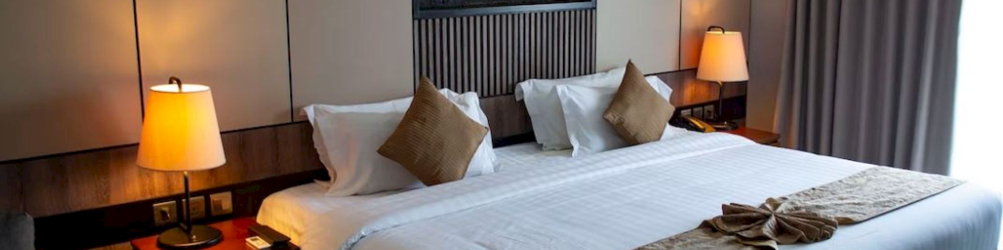 A cozy hotel room with a large bed, bedside lamps, cushions, and wall decor. A window with curtains allows in natural light from the side.