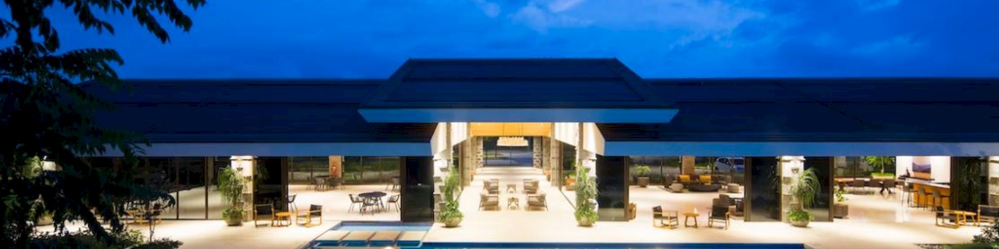 A luxurious building with an outdoor pool photographed during the evening, the area is well-lit and surrounded by lounge chairs and tables.