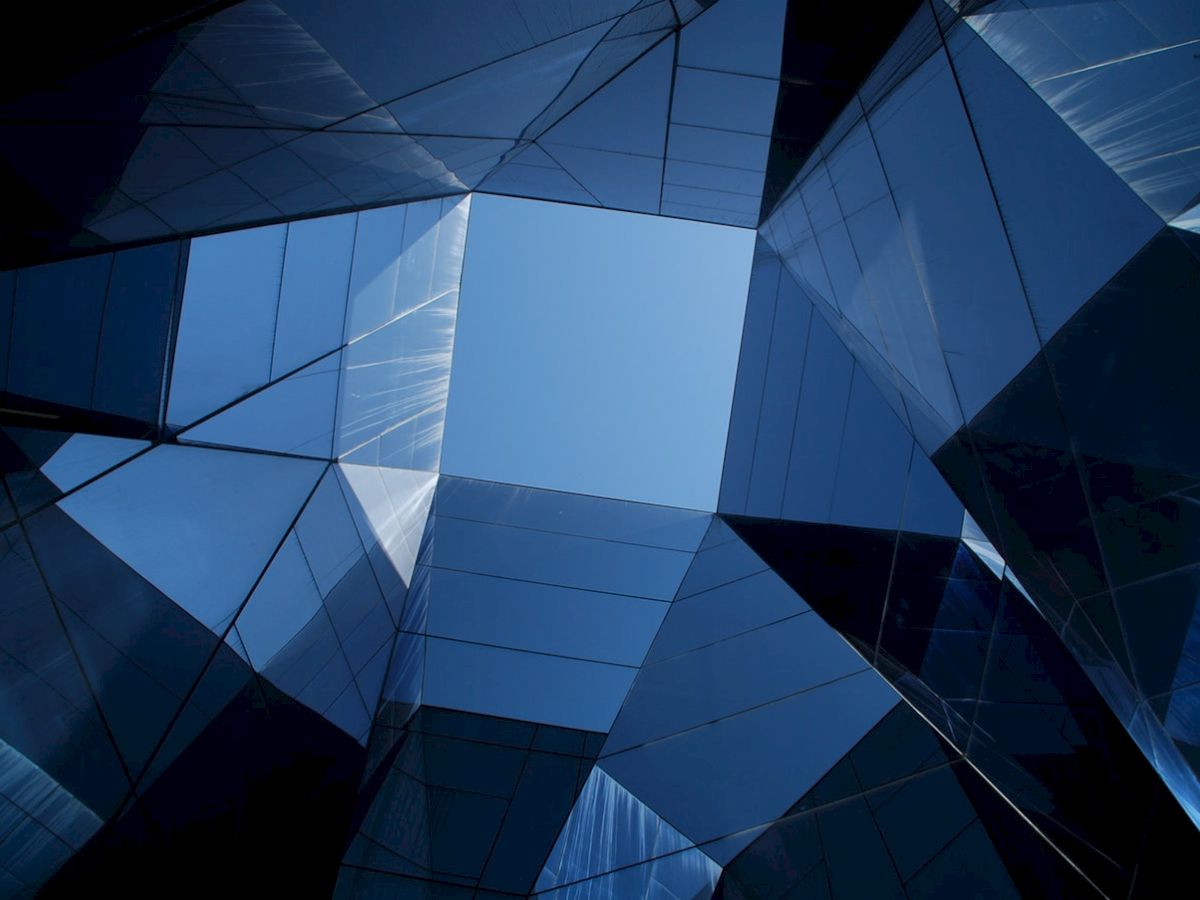 The image shows an upward view of a modern architectural structure, characterized by mirrored glass surfaces reflecting the sky.