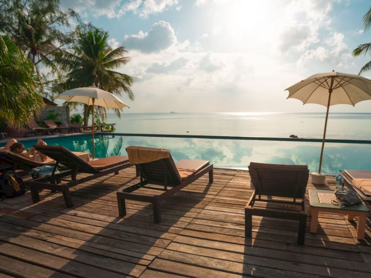 Sun loungers and umbrellas set up on a wooden deck by a pool overlooking a stunning ocean view with palm trees and a sunny sky in the background.