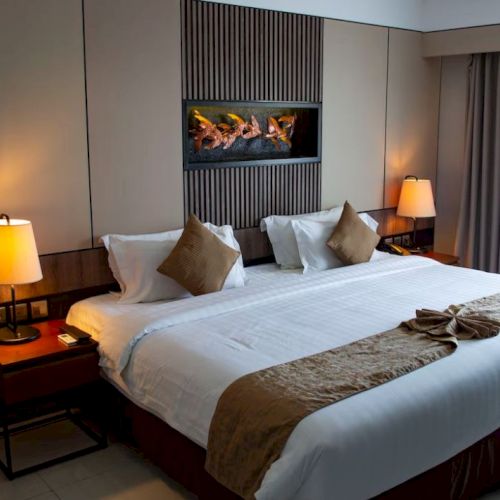 A modern hotel room features a large bed with pillows and lamps on both sides, alongside a fireplace on the wall and curtains.