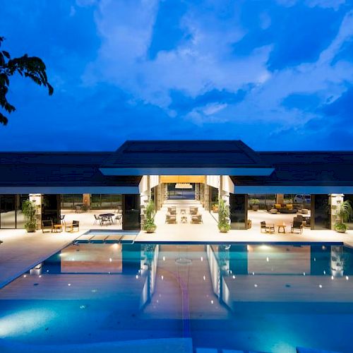 A luxurious resort with a large swimming pool, indoor seating areas, and evening lighting set under a vibrant blue sky, ending the sentence.