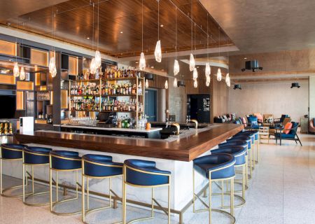 The image features a modern bar area with a wooden counter, blue bar stools, hanging lights, and a well-stocked liquor shelf in a sophisticated setting.
