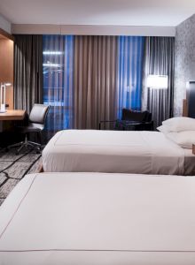 A modern hotel room with two beds, a large TV, a desk, and contemporary decor. The room features a patterned accent wall and a work area by the window.
