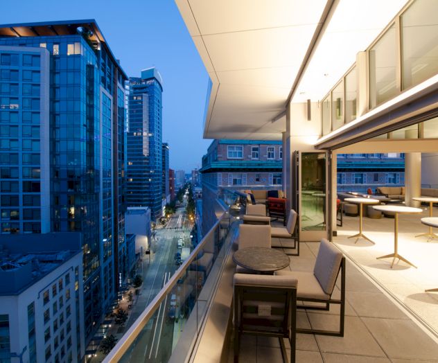 A modern city view at dusk, taken from a high balcony with seating areas and tables, overlooking high-rise buildings and a street below.