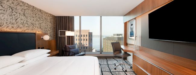 Modern hotel room with a large bed, wall-mounted TV, desk with chair, floor-to-ceiling window with city view, and contemporary decor.