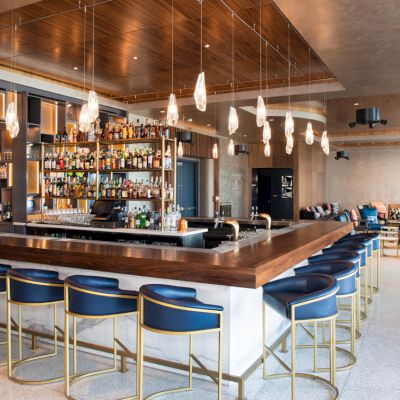 A modern bar with a wooden countertop surrounded by blue stools, decorative lights, shelves stocked with bottles, and a lounge area in the background.