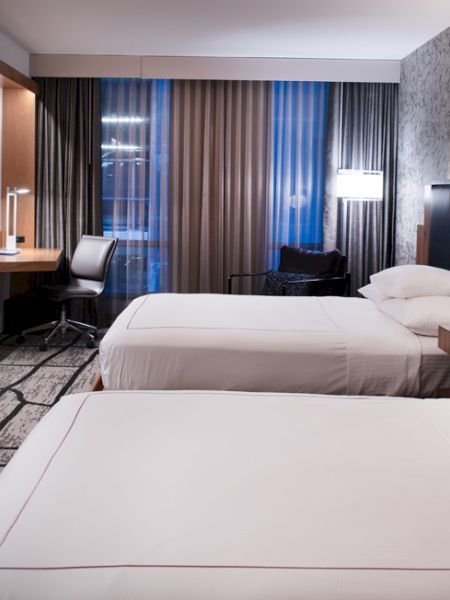 The image shows a modern hotel room with two neatly made beds, a wall-mounted TV, a desk with a chair, and stylish decor in a softly lit atmosphere.