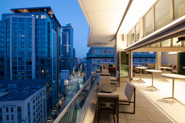 A modern urban balcony at dusk, with seating and tables overlooking a city street and high-rise buildings, lit by interior lights.