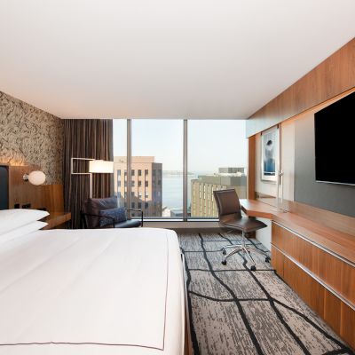 A modern hotel room with a large bed, wall-mounted TV, desk, and floor-to-ceiling window with a city view.
