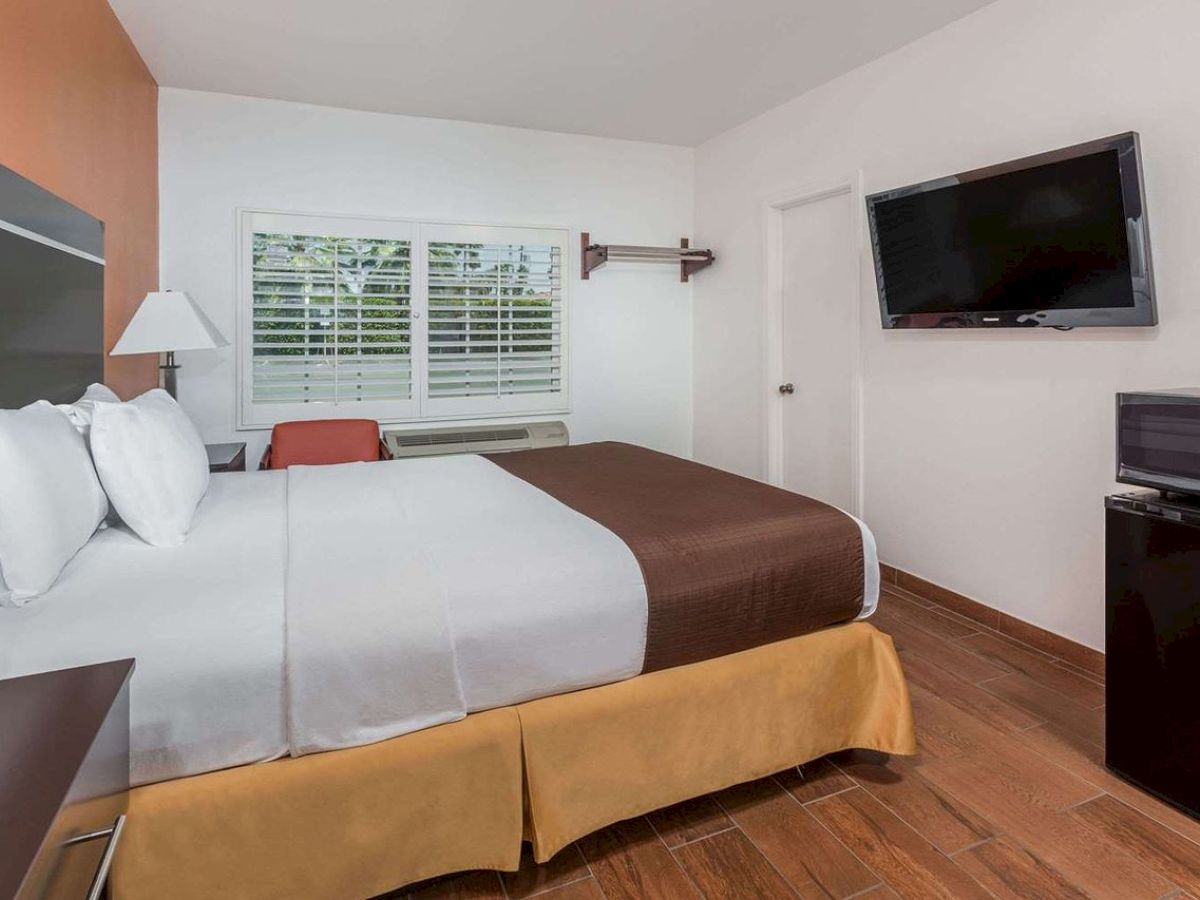 The image shows a tidy hotel room with a neatly made bed, wall-mounted TV, nightstand, window with blinds, and a small fridge.