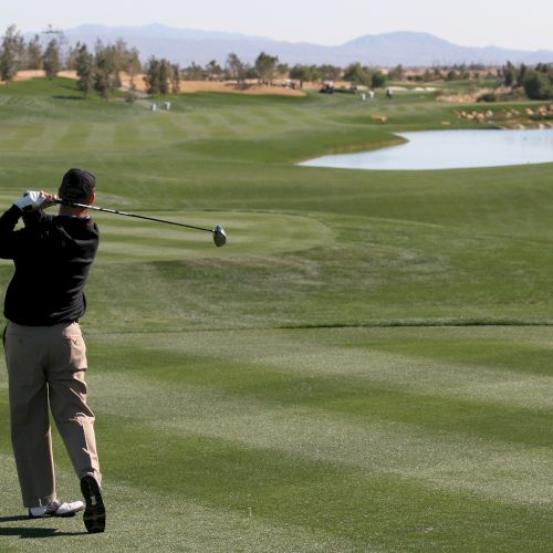 A golfer is mid-swing on a well-maintained golf course with trees and water in the background under a clear sky, creating a scenic view.