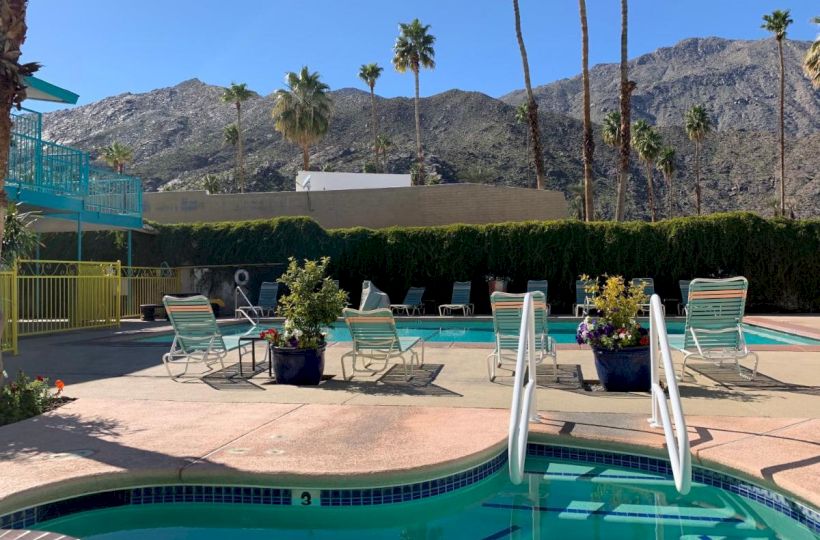 A pool area with lounge chairs, hot tub, and stairs, set against a backdrop of mountains and palm trees on a sunny day, surrounded by a green hedge.