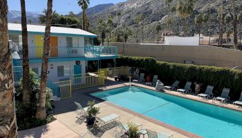 Outdoor pool area with lounge chairs, a two-story motel with colorful doors, palm trees, and mountains in the background on a sunny day.