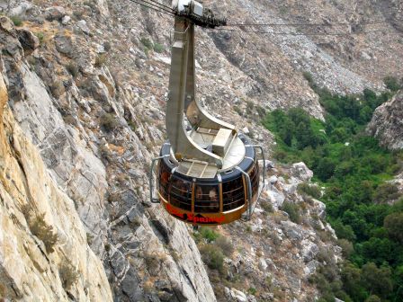 A cable car is suspended high above a rocky landscape, with green vegetation visible below. The area appears mountainous with steep rock faces.