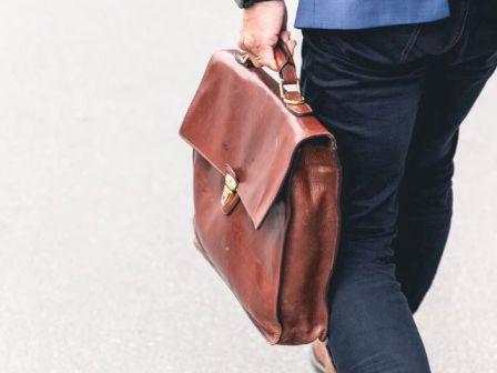 A person in a blue blazer and dark pants walks, carrying a brown leather briefcase in their right hand on a light-colored surface.