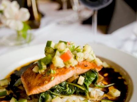 A plated gourmet dish topped with diced vegetables, served on a dark sauce, with a glass of red wine and elegant table settings in the background.