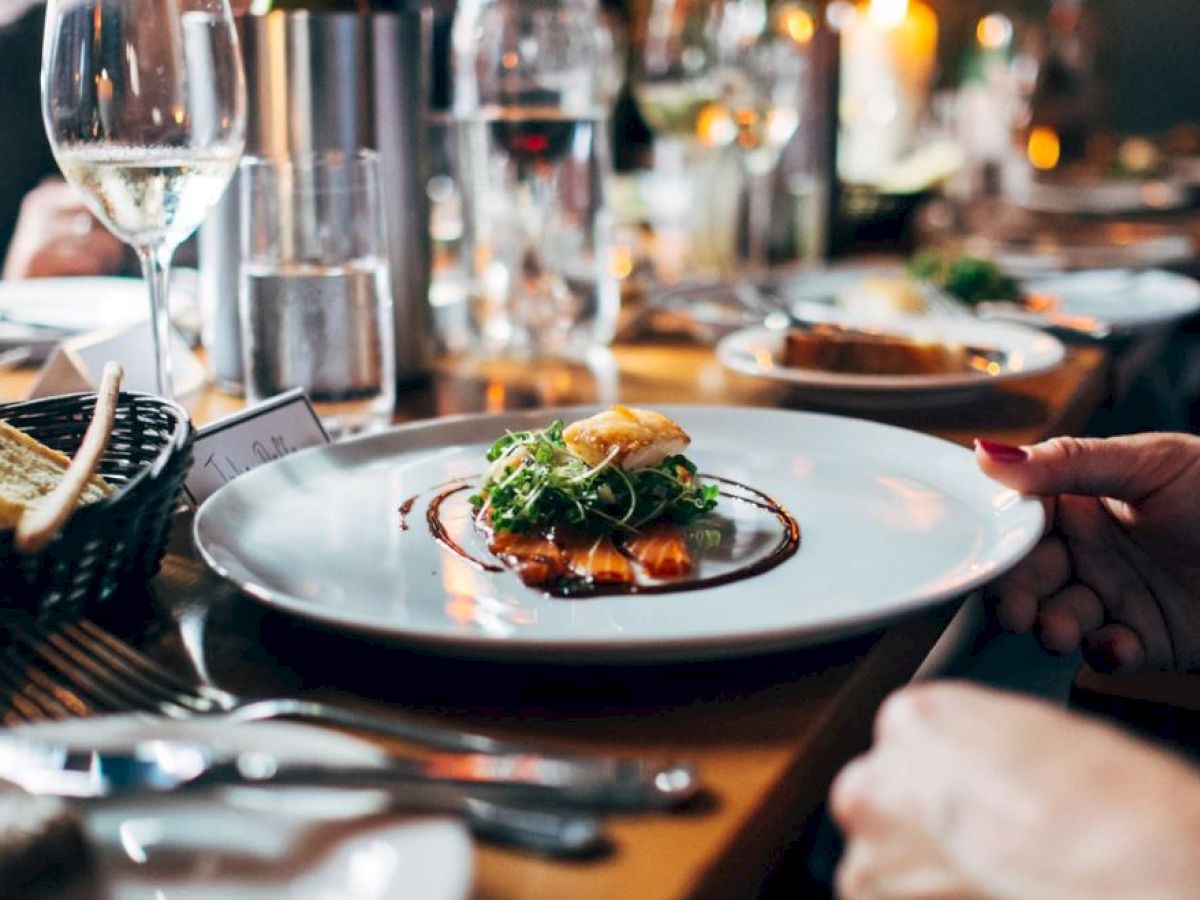 A person is holding a plate with a gourmet dish; there is bread in a basket, wine glasses, and more plates on the table, suggesting a restaurant setting.