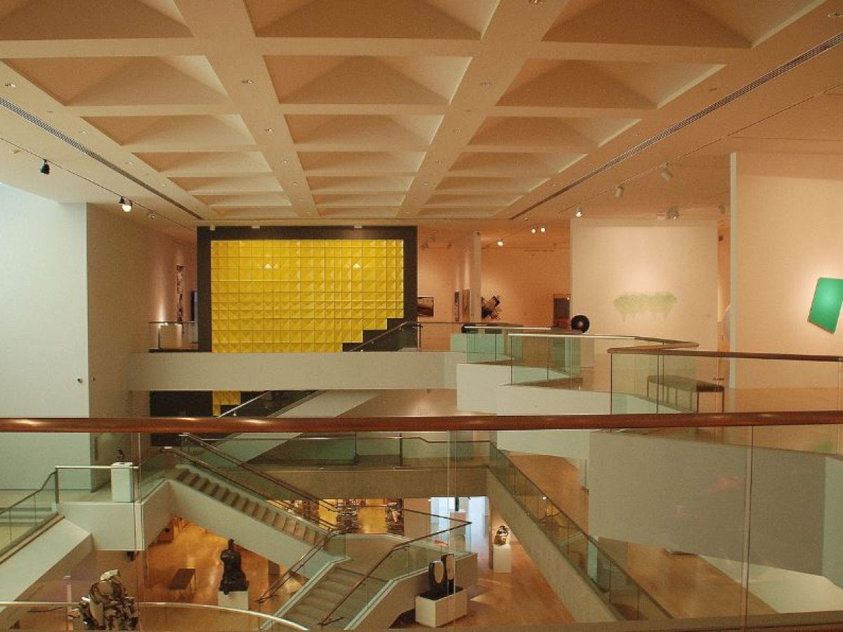 The image shows a modern art museum interior with multiple levels, abstract artworks on the walls, and a prominent yellow grid sculpture in the center.