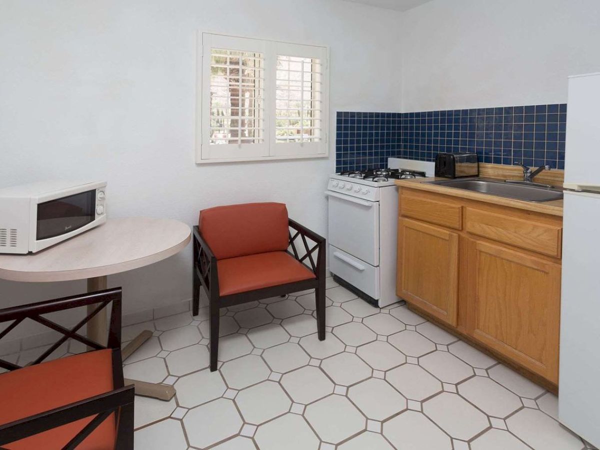 A small kitchen area with a refrigerator, stove, sink, and cabinets. It also has a round table, two chairs, and a microwave.