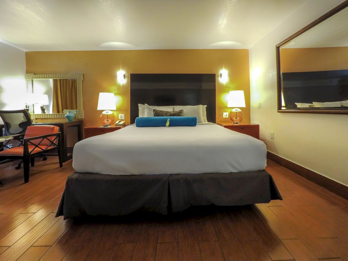 A neatly arranged hotel room features a comfortable bed with a blue bolster, bedside lamps, a large mirror, and wooden flooring, creating a cozy ambiance.