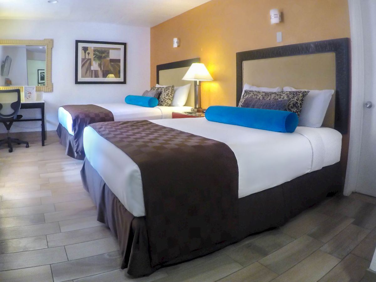 The image shows a hotel room featuring two double beds with brown and white bedding, blue bolster pillows, a desk, a chair, wall art, and a lamp.