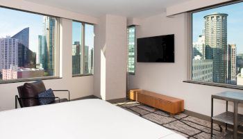 This image shows a modern hotel room with large windows, a flat-screen TV, armchair, and views of a cityscape with skyscrapers.