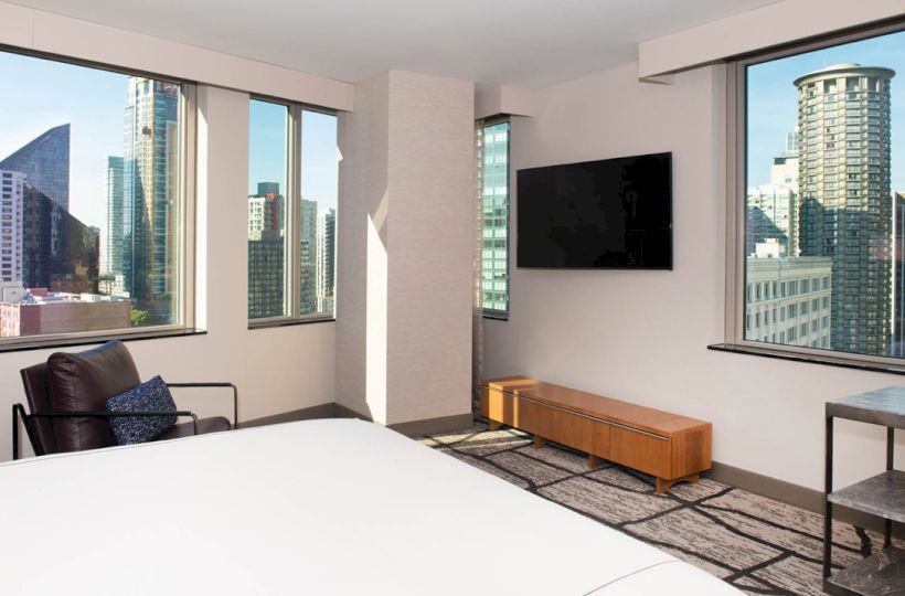 This image shows a modern hotel room with large windows, a flat-screen TV, armchair, and views of a cityscape with skyscrapers.