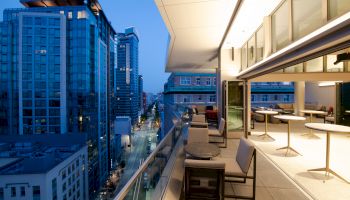 A modern cityscape at dusk with tall buildings, viewed from a stylish outdoor patio with tables and chairs on a building's balcony.