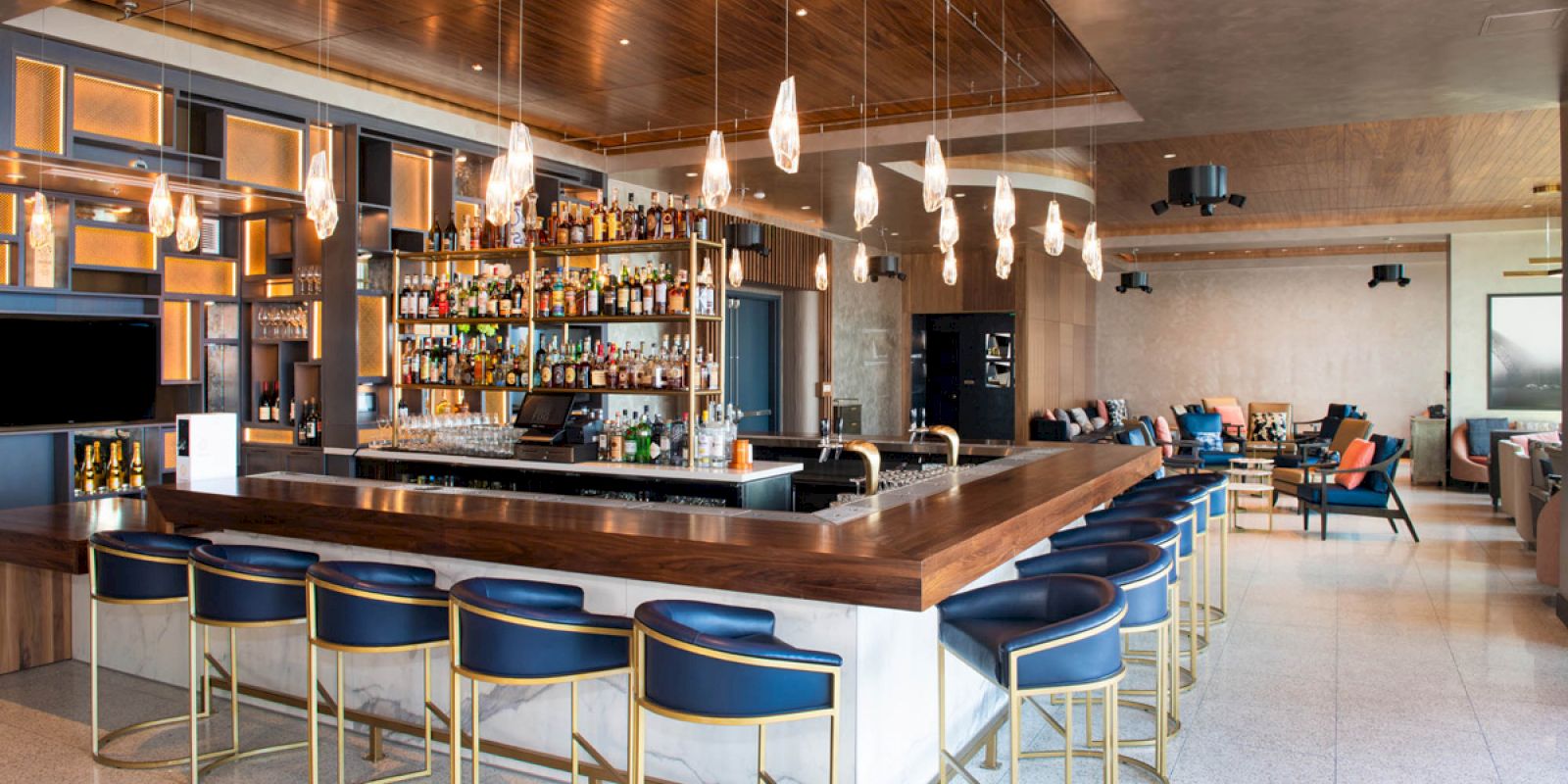 A modern bar with blue cushioned stools, a well-stocked liquor shelf, hanging lights, and comfortable seating areas.