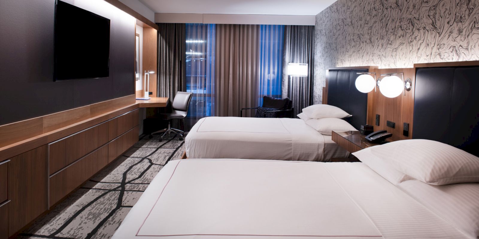 The image shows a modern hotel room with two neatly made beds, a wall-mounted TV, a desk with a chair, and contemporary decor.