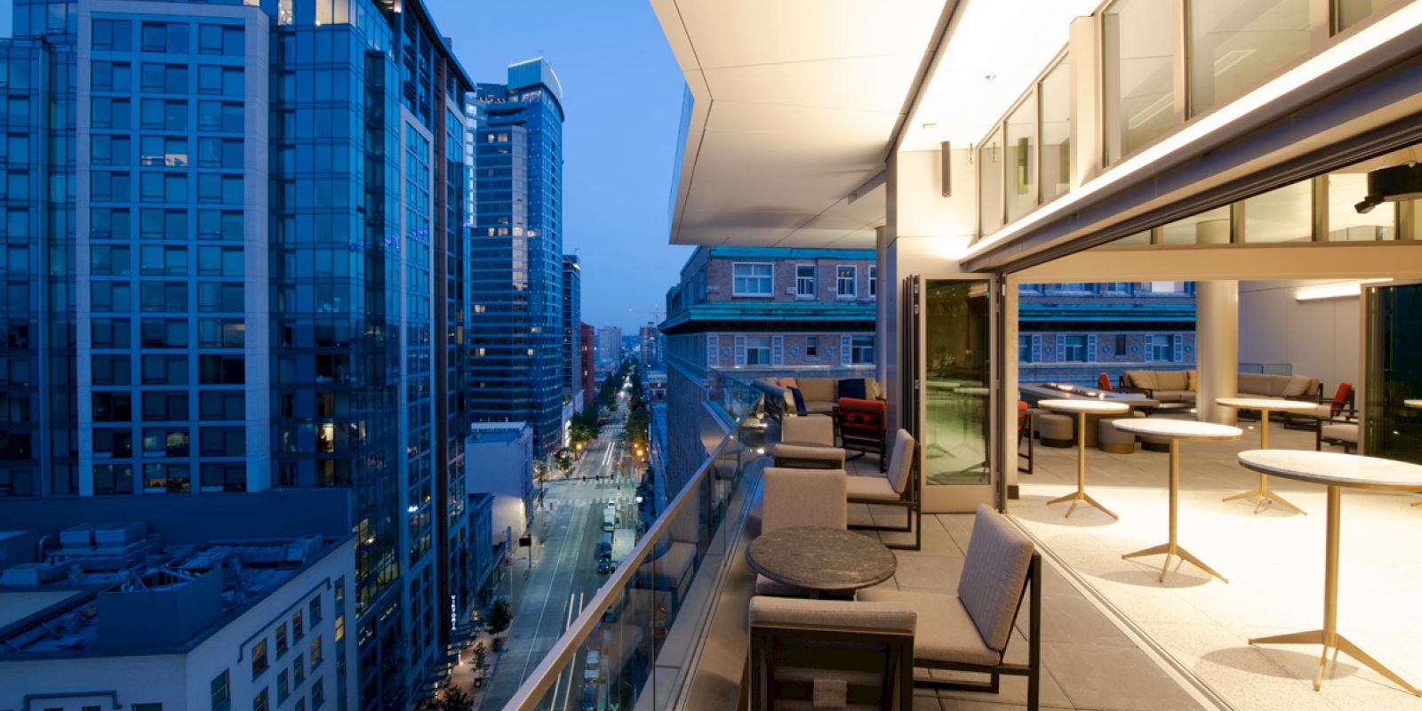 The image shows a modern, elevated terrace overlooking a cityscape with tall buildings at dusk. The terrace has tables and chairs for seating.