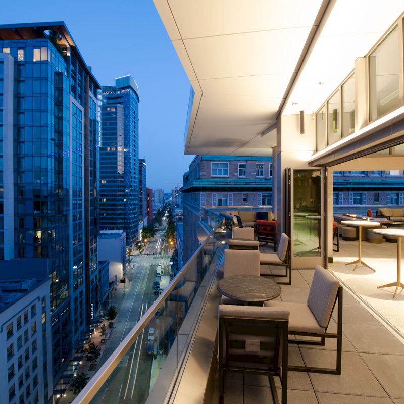 The image shows a modern rooftop patio with seating, overlooking a cityscape with tall buildings at dusk, featuring a blend of urban and cozy vibes.