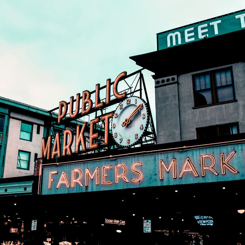 The image shows a famous public market with a large clock and neon signs reading "Public Market" and "Farmers Market" on an overcast day.