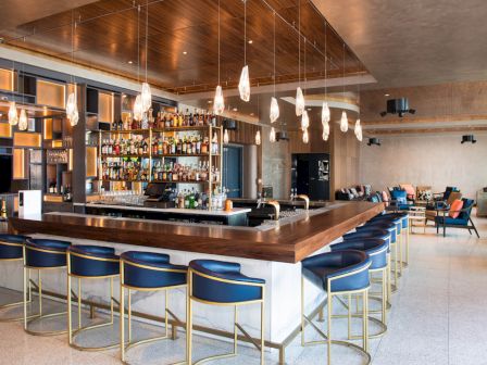 A modern bar with blue bar stools, elegant pendant lights, and a fully stocked display of liquor bottles. The seating area is visible in the background.