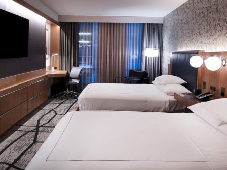 The image shows a modern hotel room with two double beds, a flat-screen TV, a desk with a chair, and a large window with curtains.