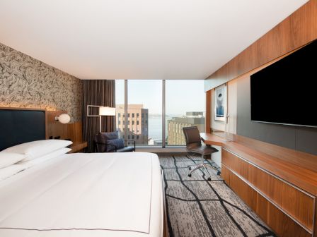 A modern hotel room with a large bed, a TV, a desk with a chair, and a large window offering a city view.