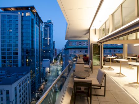 A modern balcony overlooks a city street with tall buildings, furnished with chairs and tables. The scene is lit by evening lights, creating a serene ambiance.