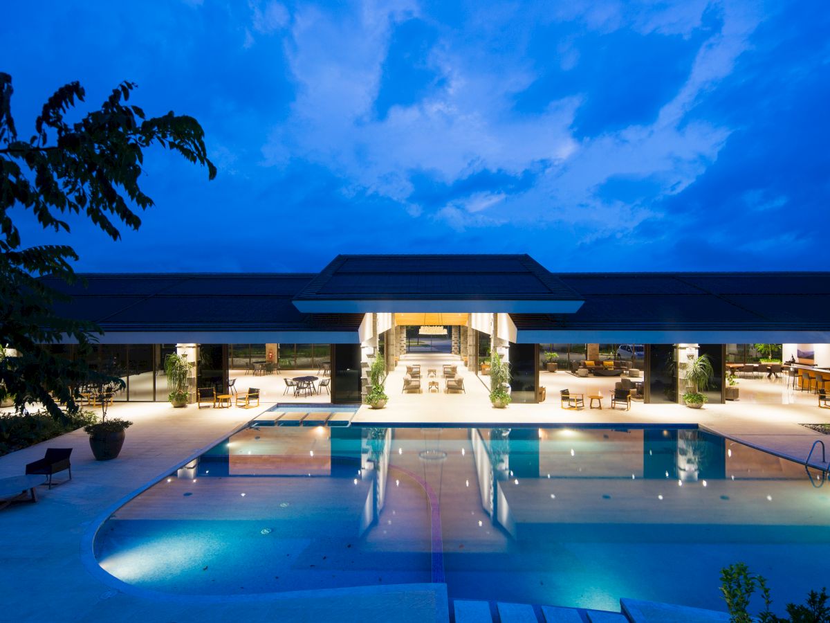 The image shows a luxurious, well-lit building with a large swimming pool in the foreground, set against an evening sky.