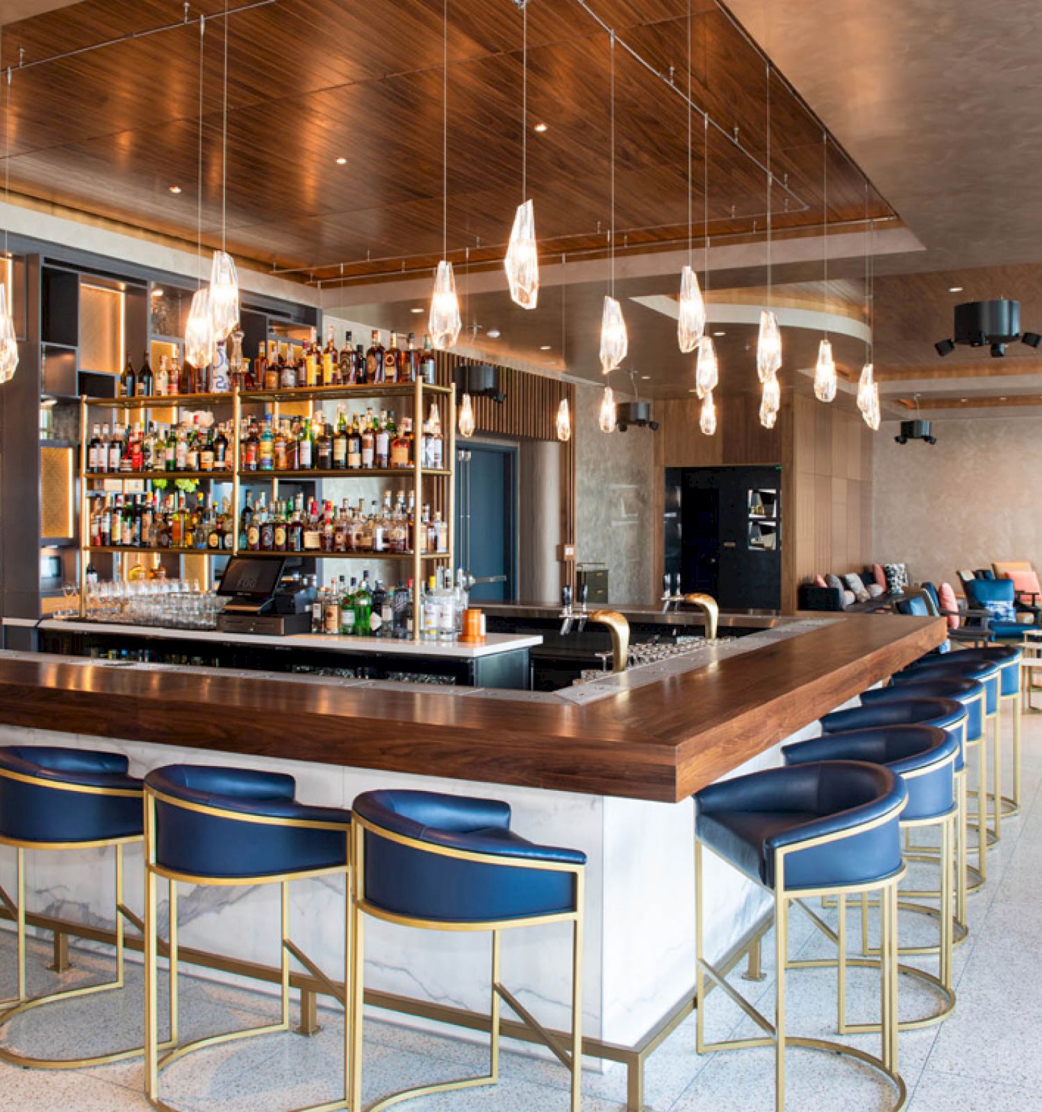 A modern bar with a wooden counter, blue chairs, pendant lights, and a well-stocked liquor shelf. The surrounding area features seating and decor.