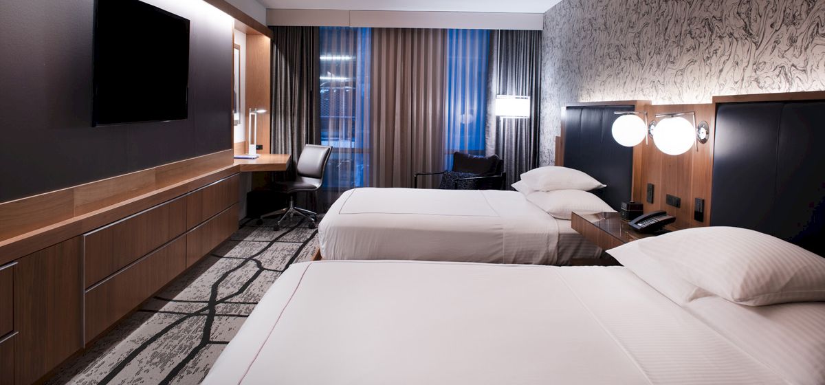 The image shows a modern hotel room with two beds, a TV, a desk, and decor elements such as curtains and wallpaper.