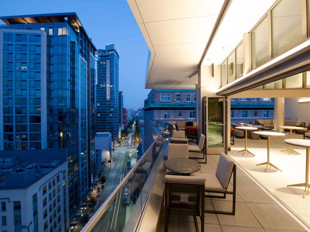 A modern cityscape view featuring tall buildings along a street at dusk, with a contemporary balcony in the foreground furnished with tables and chairs.