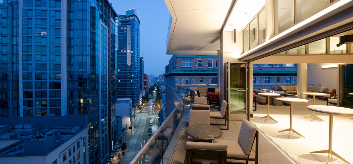 A modern cityscape view featuring tall buildings along a street at dusk, with a contemporary balcony in the foreground furnished with tables and chairs.