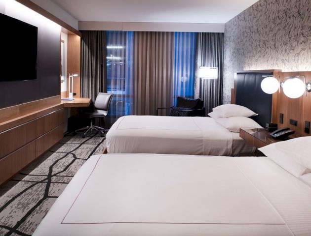 A modern hotel room with two neatly made beds, a large TV mounted on the wall, a desk with a chair, and contemporary lighting and decor ending the sentence.