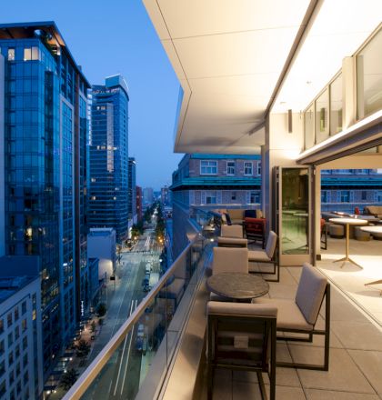 A modern cityscape viewed from a stylish balcony with seating and tables, overlooking tall buildings and a street below during twilight.