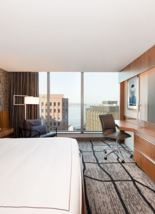 A modern hotel room featuring a large bed, a wall-mounted TV, a desk with a chair, and a window with a view of city buildings and water.